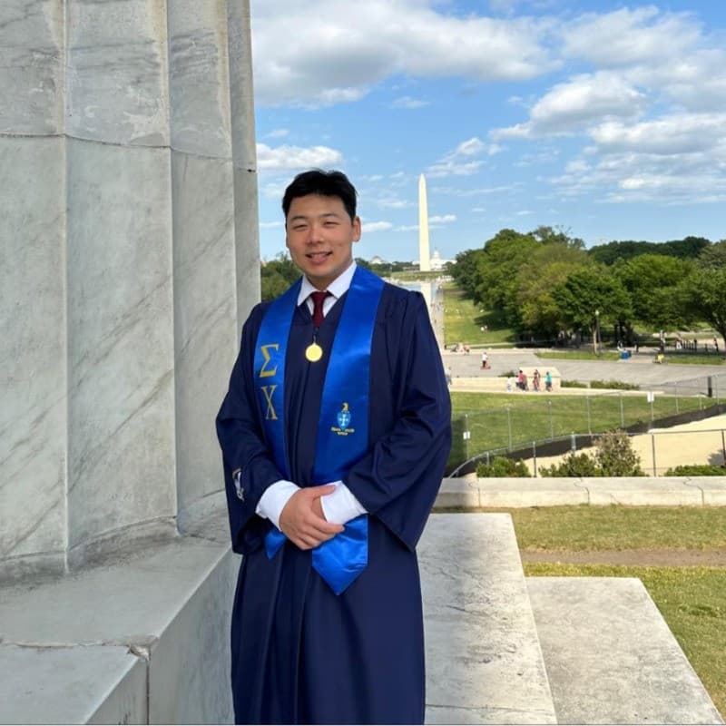 Asian graduate in blue Sigma Chi stole posing with the Washington Monument behind him.